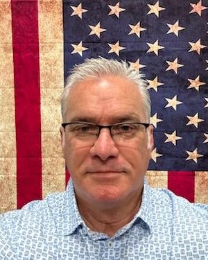 Michael Peasall, pastor of GateWay Christian Center — a man with short grey hair and dark-framed glasses in a light blue patterned button-up shirt, photographed in front of an American flag