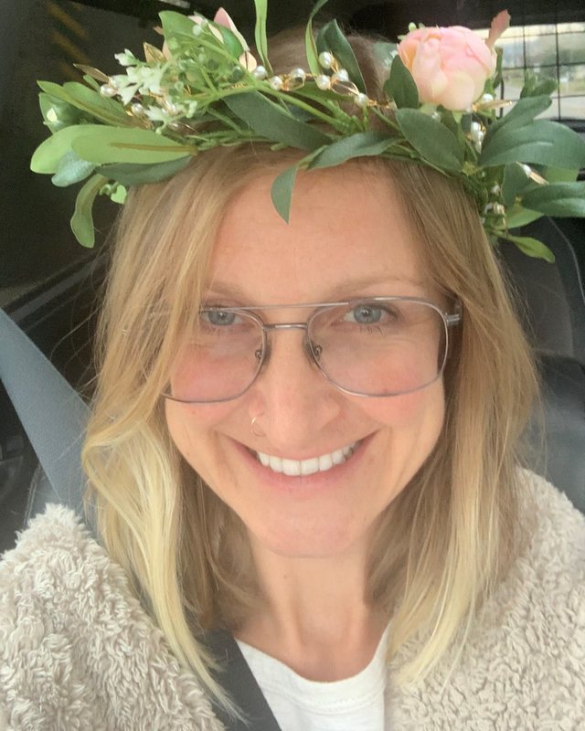 Nadine Clifton smiling — a woman with shoulder-length blonde hair and wire-framed glasses wearing a crown of greenery and pale pink flowers