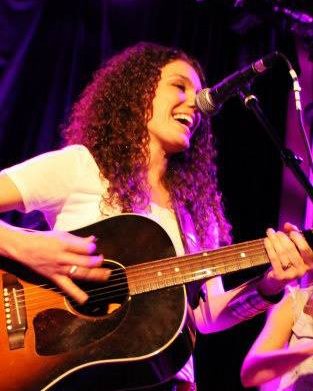 Sarah McGuffey singing into a microphone while playing an acoustic guitar under warm stage lighting